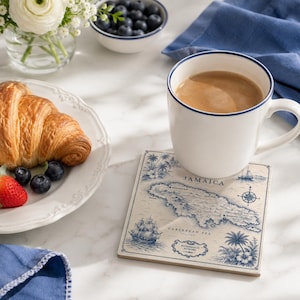 May include: A square ceramic coaster featuring a vintage-style blue and cream map of Jamaica, complete with illustrations of a sailing ship, palm trees, and a compass rose. The coaster sits beneath a white ceramic mug filled with coffee.