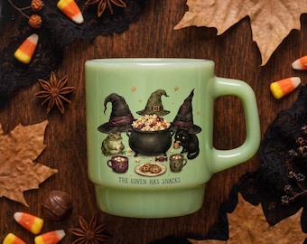 May include: A light green mug featuring a graphic illustration of three witch hats, a black cauldron filled with snacks, a frog, a black cat, two small mugs, and cookies. The text THE COVEN HAS SNACKS is printed in black below the illustration.
