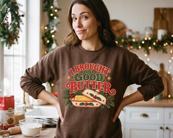 May include: A dark brown long-sleeve sweatshirt featuring a graphic print of a butter dish with the text I BROUGHT THE GOOD BUTTER in red and green lettering, surrounded by festive pine branches and red holly berries on the front center.