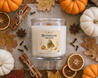 May include: A clear glass candle jar filled with white wax, featuring a label that reads Mushroom Mood with an illustration of a frog on a mushroom. The candle is surrounded by orange and white pumpkins, cinnamon sticks, dried orange slices, and leaves.