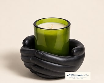 May include: A green glass candle in a matte black holder shaped like two cupped hands. The candle features a single white wick. A small white rectangular logo in the bottom right corner displays the text Q&WLT QUIRE & WEFT PAPER SCENT STORE.