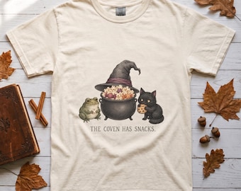 May include: A cream-colored short-sleeve t-shirt featuring a graphic of a frog and a black cat sitting next to a black cauldron filled with popcorn. A witch hat sits atop the cauldron. The text below reads, THE COVEN HAS SNACKS.