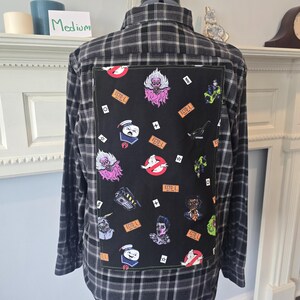 Può includere: Una camicia di flanella a quadri neri e grigi di medie dimensioni, con una toppa quadrata nera sul retro. La toppa presenta immagini di cartoni animati colorati e il testo "ECTO-1". La camicia è esposta su un manichino.
