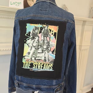 May include: A blue denim jacket with a black patch featuring a colorful graphic of a group of people and the text "DON'T CROSS THE STREAMS". The jacket is displayed on a mannequin.