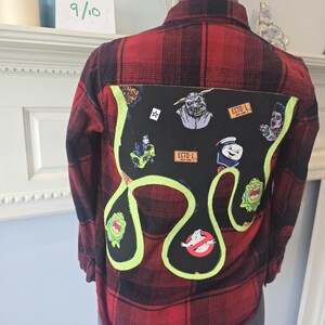 Può includere: Camicia di flanella a quadri rossi e neri con una toppa nera con personaggi di Ghostbusters e il logo Ghostbusters. La toppa è bordata di verde neon. La camicia è esposta su un manichino.