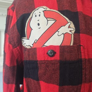 Può includere: Camicia a quadri rossi e neri con una toppa con il logo Ghostbusters sul petto. Il logo presenta un fantasma bianco all'interno di un cerchio rosso con una linea diagonale rossa. La camicia ha una tasca singola con un bottone nero.