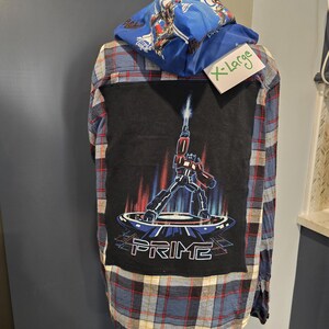 Peut inclure: Chemise à carreaux bleue, rouge et noire avec un motif noir au dos représentant un robot et le mot "PRIME". Une capuche bleue avec un motif de robot est attachée. La chemise est étiquetée "X-Large".