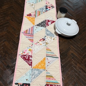 May include: A long, rectangular quilted table runner featuring a patchwork design of various fabric triangles in floral, striped, and solid patterns. The runner has a solid light beige background with a pink border and visible diagonal stitching.