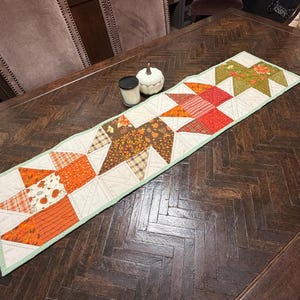 May include: A rectangular quilted table runner featuring a patchwork design of autumn-themed fabrics in shades of orange, brown, green, and cream. The runner displays geometric star patterns with leaf, pumpkin, and plaid prints, finished with a light green border.