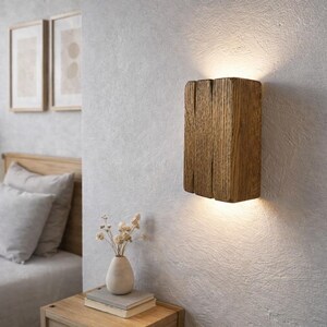 Può includere: Una lampada da parete rettangolare realizzata con un pezzo massiccio di legno marrone scuro testurizzato. L'apparecchio è montato verticalmente su una parete ed emette una luce calda e soffusa sia dal bordo superiore che da quello inferiore.