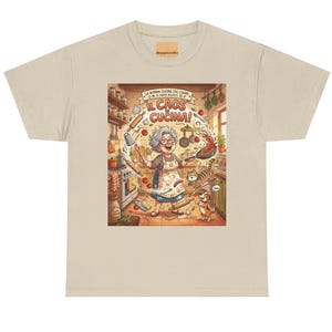 Può includere: T-shirt beige con un'illustrazione colorata di una scena di cucina. L'opera raffigura una donna anziana con un grembiule, circondata da cibo e utensili da cucina volanti. Il testo sulla maglietta recita "IL CAOS CUCINA!" e "LA NONNA CUCINA COL CUORE".