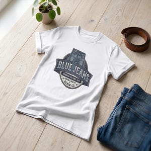 Può includere: Maglietta bianca con grafica "Blue Jeans", un paio di jeans blu e una cintura in pelle marrone su una superficie di legno. La maglietta presenta un logo a tema denim con la scritta "Handcrafted since 1978". È visibile anche una piccola pianta in vaso.