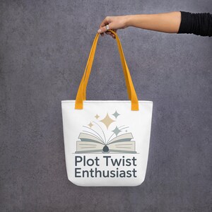 May include: White tote bag with yellow handles. The bag features an open book graphic with stars and the text "Plot Twist Enthusiast." The tote bag is held up against a grey background. The bag is designed for carrying books and other items.