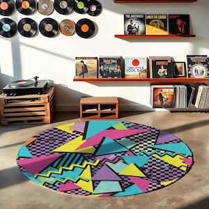 Op de afbeelding: Een ronde vloerkleed met een kleurrijk jaren 80 design. Het heeft driehoeken, zigzags en dambordpatronen in roze, geel, blauw en paars. Een platenspeler staat op een houten krat.