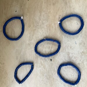 May include: Five blue disc bracelets are arranged on a light brown surface. Each bracelet is a circle of small, flat, blue discs strung together. White string is visible at the ends of each bracelet.