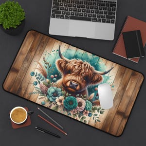 May include: A rectangular desk mat featuring a Highland cow illustration with a floral arrangement. The design includes a brown cow with long horns, surrounded by teal and blue flowers, set against a wooden plank background.