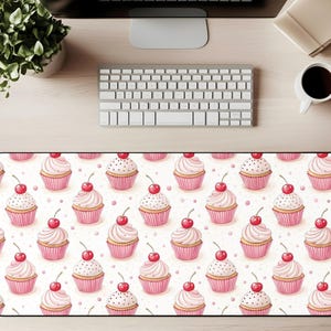 May include: A rectangular desk mat featuring a repeating pattern of pink cupcakes with white frosting and a red cherry on top. The cupcakes are set against a white background with small pink dots. The mat is surrounded by a black border.