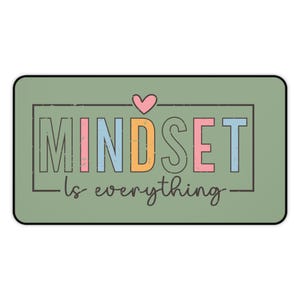 以下が含まれることがあります： 「MINDSET is everything」という言葉が書かれた、長方形のセージグリーンのデスクマット。 「MINDSET」という単語は、ピンク、オレンジ、ピンク、ブルー、ピンクと、文字ごとに異なる色でボックスに入っています。 「I」の上にはピンクのハートがあります。