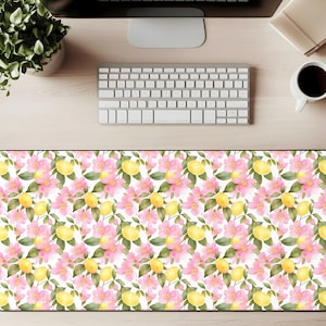 May include: A rectangular desk mat featuring a watercolor pattern of yellow lemons, pink flowers, and green leaves on a white background. The mat is surrounded by a black border. The desk mat is on a light wood surface.