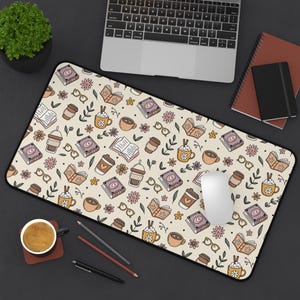 May include: A rectangular beige mouse pad with a black border, covered in a repeating pattern of books, coffee cups, flowers, and eyeglasses. A laptop, notebooks, and a coffee cup are on the dark surface.