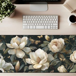 May include: A rectangular desk mat featuring a floral design with large white magnolia flowers, butterflies, and buds against a dark background. The mat has a black border and is placed on a wooden desk.