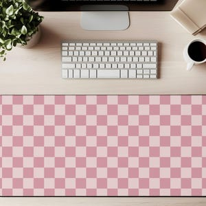 May include: A pink and light pink chequered pattern mouse mat. The rectangular mat is on a light wood desk with a keyboard, monitor, coffee cup, and a small plant in a white pot. The mat has a black border.