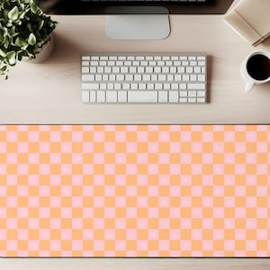 Puede incluir: Una alfombrilla de escritorio rectangular con un borde negro y un patrón repetitivo de cuadrados naranjas y rosas. La alfombrilla está diseñada para proteger la superficie de un escritorio y proporcionar una superficie lisa para un ratón y un teclado. La alfombrilla se muestra en un escritorio de madera clara.