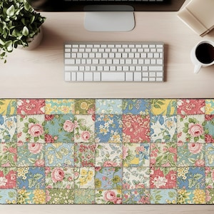 Puede incluir: Una alfombrilla de escritorio rectangular con un diseño de patchwork con estampados florales en tonos rosa, azul, amarillo y verde. La alfombrilla está rodeada por un borde negro y se encuentra sobre un escritorio de madera de color claro. También se ven un teclado, un monitor y una taza de café.
