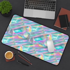 May include: A rectangular desk mat with a black border features a repeating pattern of iridescent, translucent shapes. A white computer mouse rests on the mat. A laptop, notebook, and coffee cup are also visible.