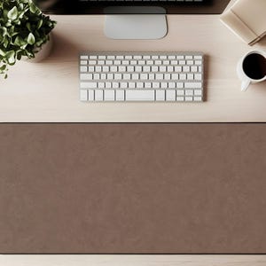 May include: A brown rectangular desk mat with rounded corners. The mat is made of a textured material and has a black border. A white keyboard, a cup of coffee, and a small plant are visible on the desk.