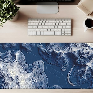 May include: A large, rectangular desk mat with a dark blue topographical design. The design features white contour lines that create a 3D effect, resembling a map. The mat is placed on a light-colored wooden desk, with a keyboard, monitor, and coffee cup visible.