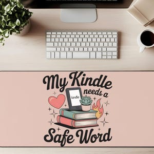 May include: A rectangular desk mat with a pink background featuring the text "My Kindle needs a Safe Word." The design includes a Kindle, books, a heart, and a small plant. A keyboard, monitor, and coffee cup are visible.