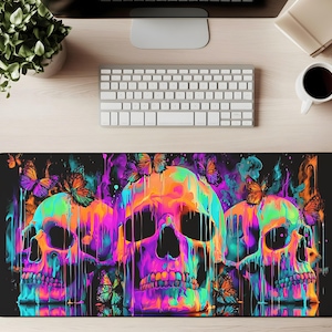 Puede incluir: Una alfombrilla de escritorio negra con tres calaveras coloridas con efectos de pintura goteante en tonos naranja, rosa, morado y verde azulado. Hay mariposas dispersas alrededor de las calaveras. La alfombrilla de escritorio es rectangular y está sobre un escritorio de madera clara.