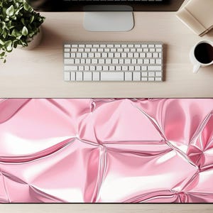 May include: A large rectangular pink desk mat with a shiny, crumpled foil design. The mat is on a light wood desk, with a keyboard, monitor, coffee cup, and plant visible.
