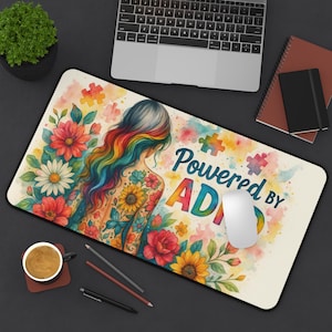 May include: A rectangular mouse mat with a vibrant design. It depicts a woman with rainbow hair and floral tattoos, surrounded by flowers and the text "Powered by ADHD". A computer mouse sits on the mat.