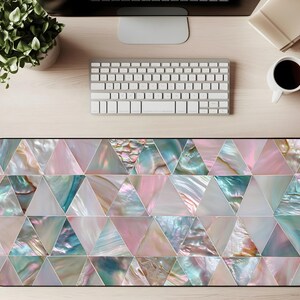 May include: A rectangular desk mat with a geometric design of iridescent triangles in shades of pink, teal, and white. The mat is placed on a light wood desk, with a keyboard, monitor, and coffee cup visible.