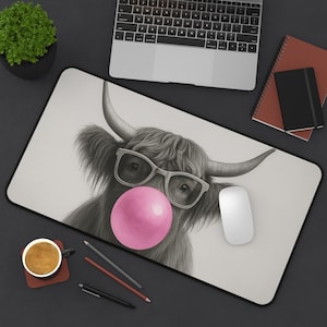 May include: A rectangular mouse pad featuring a black and white illustration of a Highland cow wearing glasses and blowing a pink bubble. The mouse pad has a black border and is surrounded by office supplies.