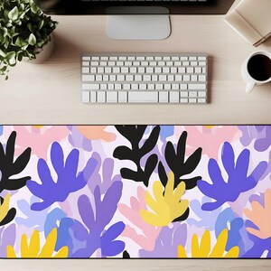 Pode incluir: Um tapete de mesa retangular com um design floral abstrato colorido. O padrão inclui formas de folhas pretas, azuis, amarelas, rosa e roxas sobre um fundo branco. O tapete está colocado numa secretária de madeira clara, com um teclado, um monitor e uma chávena de café.