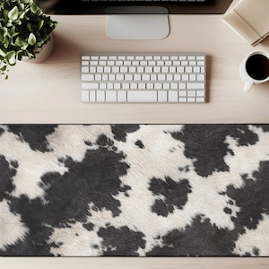 May include: A rectangular desk mat with a cowhide pattern in black and white. The mat is placed on a light wood desk, with a keyboard, monitor, and coffee cup visible in the background.