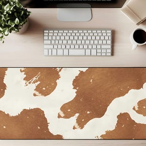 Pode incluir: Tapete de mesa retangular com um padrão de pele de vaca marrom e branco. O tapete tem uma borda preta. Um teclado de computador, um monitor, uma xícara de café e uma planta estão em uma mesa de madeira clara.