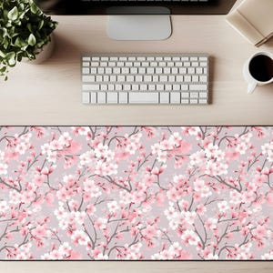 May include: A rectangular desk mat with a floral design of pink cherry blossoms on a light grey background. The mat has a black border and is designed to protect the desk surface from scratches.