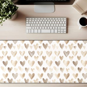 Puede incluir: Una alfombrilla de escritorio rectangular con un patrón repetido de corazones de acuarela en tonos beige y gris sobre un fondo blanco. La alfombrilla está diseñada para proteger la superficie de un escritorio y proporcionar un área lisa para un teclado y un ratón.