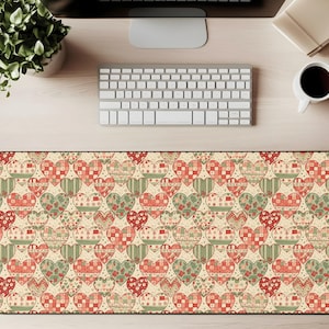 Puede incluir: Alfombrilla de escritorio rectangular con un patrón repetido de corazones en tonos rojos, verdes y beige. Los corazones tienen varios diseños, incluyendo rayas y patchwork, sobre un fondo crema. La alfombrilla está colocada sobre un escritorio de madera clara.