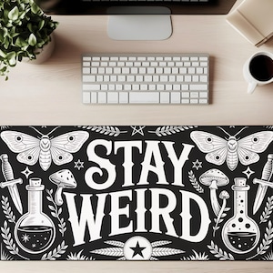 May include: Black rectangular desk mat with a white graphic design. The design includes two moths, mushrooms, potion bottles, daggers, stars, and the words "STAY WEIRD" in bold letters. The mat is surrounded by a white keyboard, a coffee cup, and a plant.