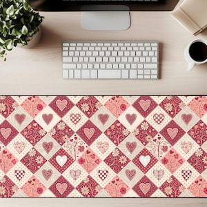 Puede incluir: Alfombrilla de escritorio rectangular con un patrón repetitivo de corazones en rojo, rosa y blanco. El diseño se asienta sobre un fondo crema y burdeos, creando un efecto de patchwork. La alfombrilla está diseñada para colocarse debajo de un teclado y un ratón.