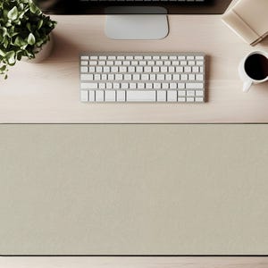 May include: A top-down view of a desk setup featuring a beige rectangular desk mat with a black border. A white keyboard, a computer monitor, a cup of coffee, a notepad, and a potted plant are also visible on the light wood desk.