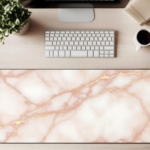 May include: A rectangular desk mat with a marble design. The mat features a white and blush pink background with gold veining. A black border frames the mat. The mat is on a light wood surface, with a keyboard, monitor, and coffee cup visible.