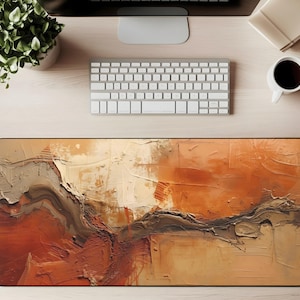 May include: An abstract art mouse pad with a black border. The design features a textured, layered composition in shades of orange, brown, and beige, resembling a landscape or geological formation. The mouse pad is rectangular and sits on a light wood desk.