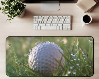 May include: A golf ball with water droplets rests in green grass, creating a macro shot. The golf ball is white with dimples. The image is on a desk mat, with a keyboard, monitor, coffee cup, and plant in the background.