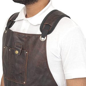 May include: A brown leather apron with a pocket and metal rivets. The apron has black straps and padded shoulder straps. The apron is worn over a white collared shirt.
